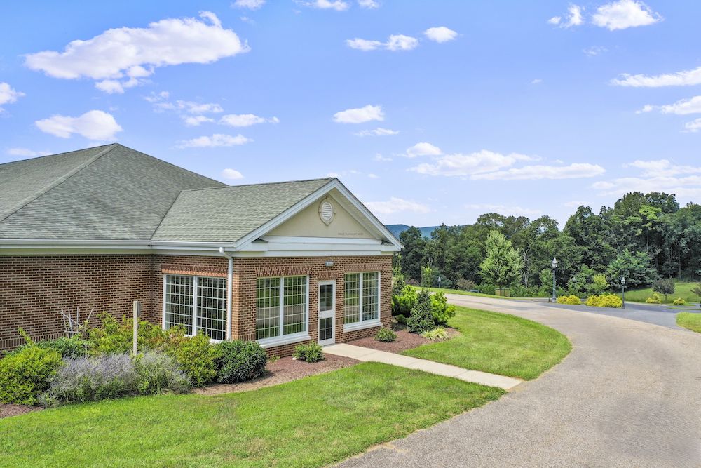 Assisted Living 3 A brick building with large windows stands amid greenery, overlooking a winding path and scenic landscape under a sunny, blue sky.