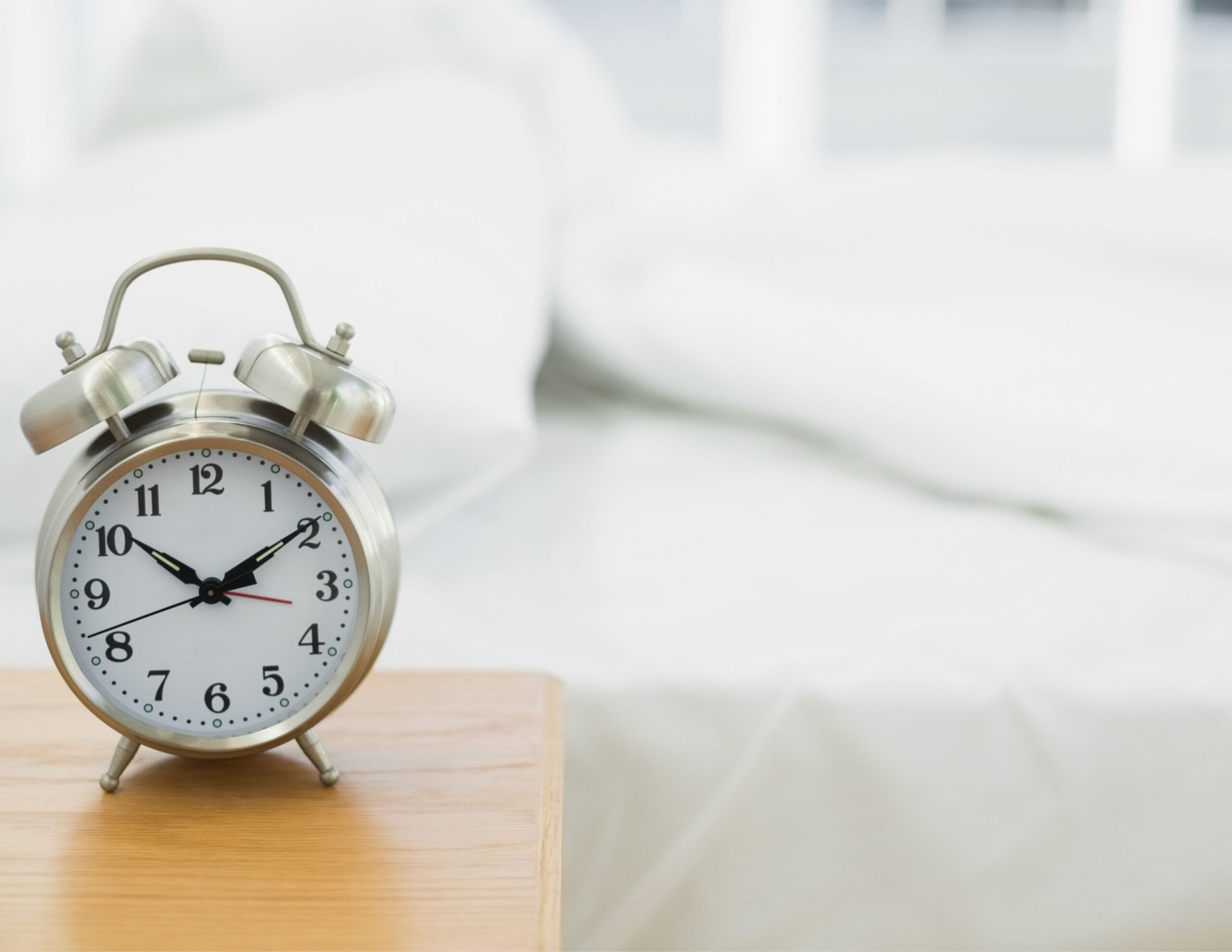 Home 5 A silver alarm clock sits on a wooden bedside table next to a neatly made bed with white sheets in a bright room.