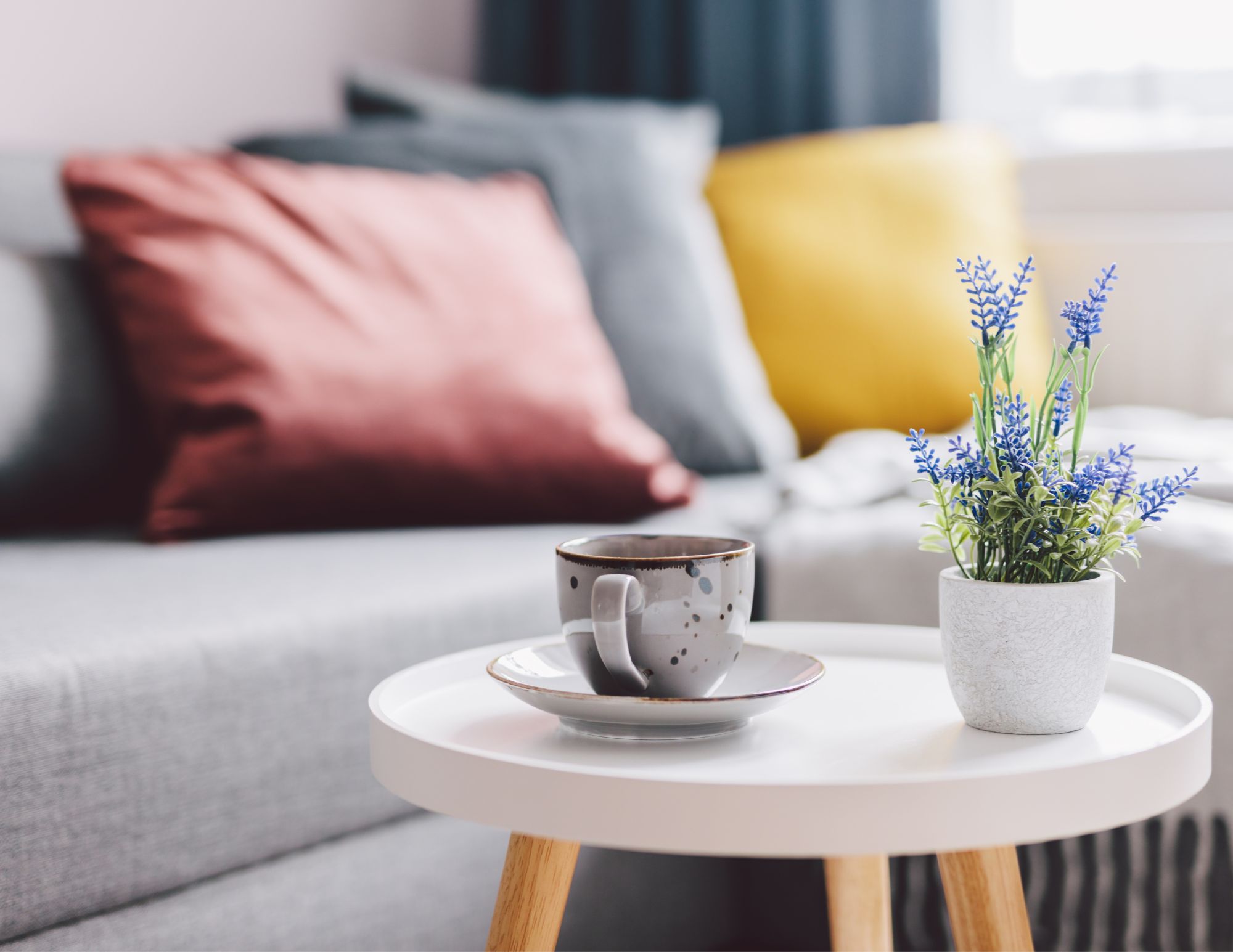 Home 3 A cozy living room scene with a white round table, gray couch, colorful cushions, coffee mug, and a small potted lavender plant.