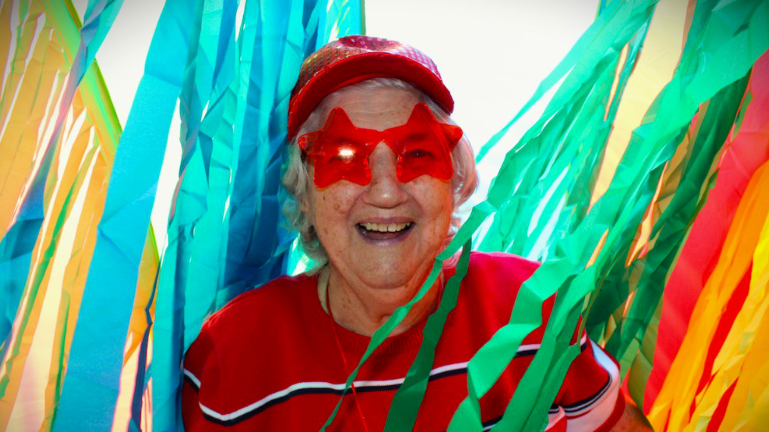 A smiling person with star-shaped sunglasses stands among colorful streamers, wearing a red cap and sweater, creating a festive atmosphere.