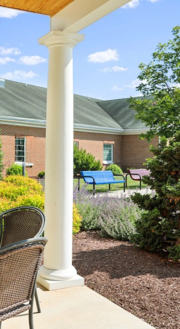 A sunny courtyard features brick building, column, wicker chair, colorful benches, and neatly trimmed bushes under a clear blue sky.