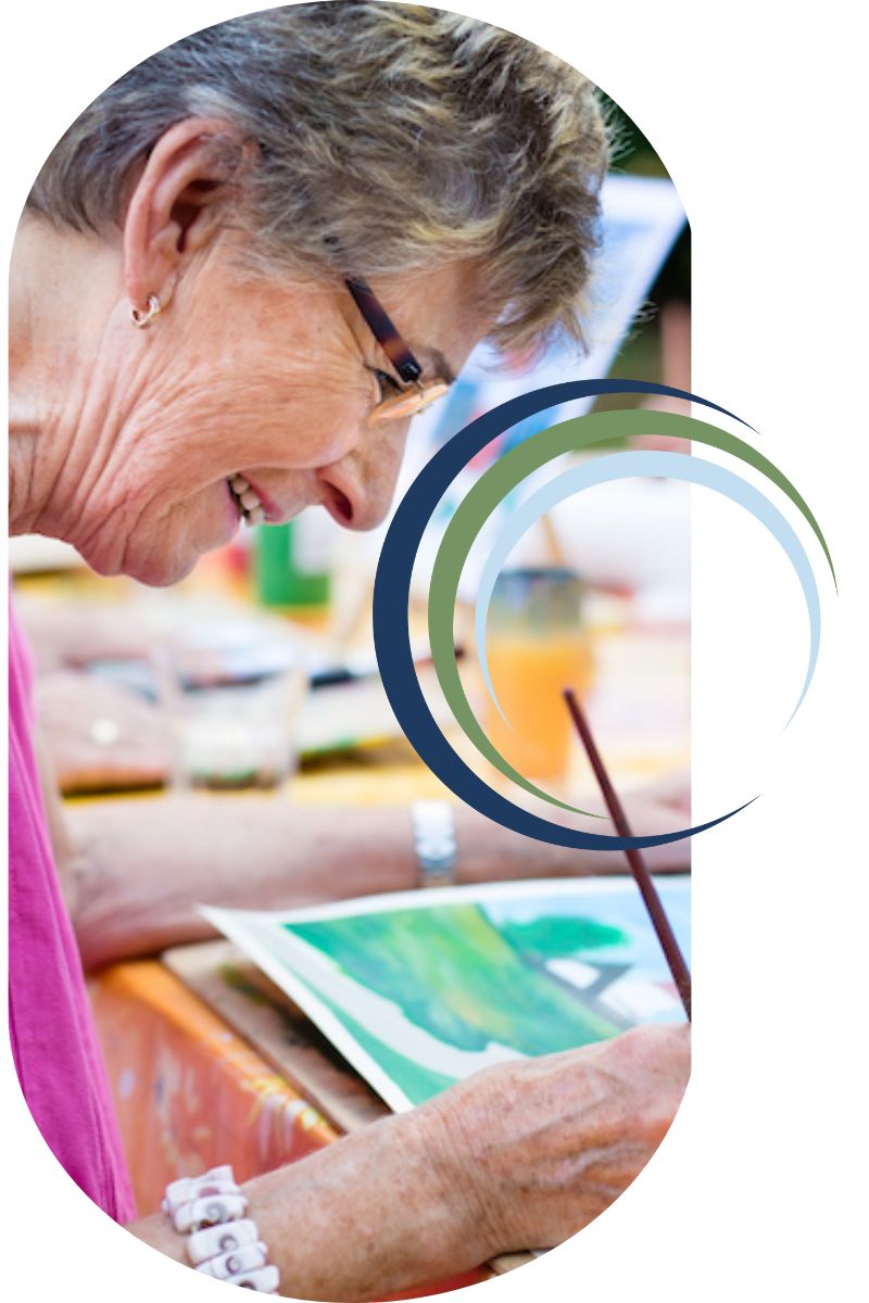 A person wearing glasses is joyfully painting at a table, focusing on a colorful canvas, with vibrant surroundings visible.