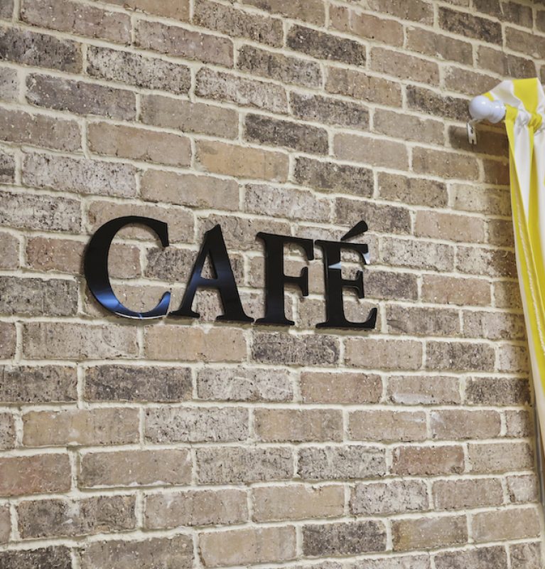 Brick wall with "CAFÉ" sign and yellow-striped curtain. Bright, inviting atmosphere suggesting a cozy café setting.