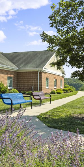 A brick building with benches on a path surrounded by trees and flowers under a clear blue sky.
