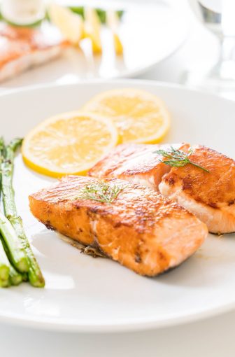 Grilled salmon fillets garnished with dill sit beside asparagus and lemon slices on a white plate, creating a fresh, vibrant dish.