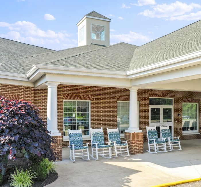 A brick building with white columns and rocking chairs on the porch, surrounded by neatly landscaped greenery under a clear blue sky.