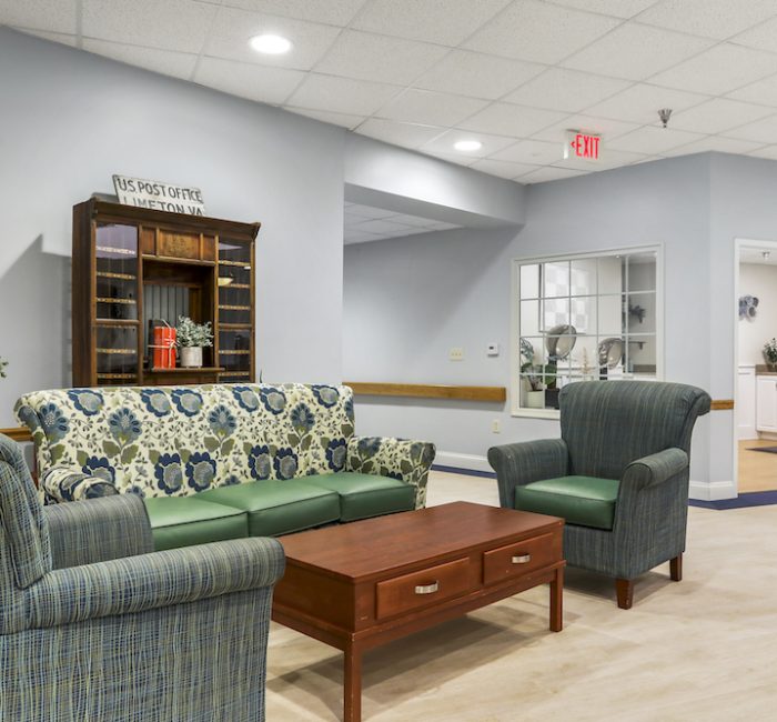 Room with floral-patterned sofa, armchairs, wooden table, US Post Office cabinet, and potted plants. Bright lighting and neutral-toned walls.