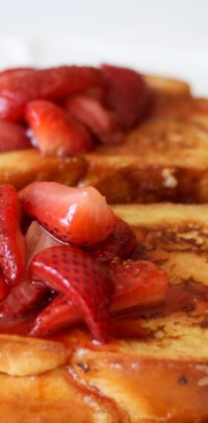 Two slices of French toast topped with fresh, juicy strawberries on a plate. The toast is golden and drenched in syrup.