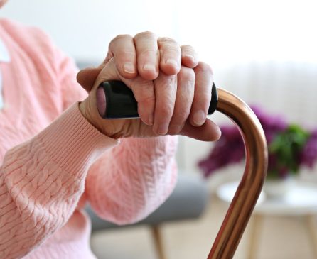 A person with folded hands rests on a cane. They wear a pink sweater. Blurred lilacs in the background suggest a cozy setting.